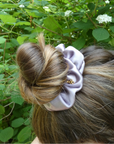 A lavender satin scrunchie with a small gold bead accent, holding up a brown and blonde high bun hairstyle outdoors against a background of green leaves and white flowers.