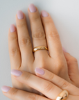 A hand wearing a gold dome ring with light pink nail polish on a neutral background.