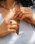 Close-up of a person wearing a stack of gold rings and a layering of necklaces with blue sodalite beads and pebble charm. / belue bijoux