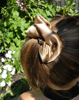 A large satin hair bow accessory in a champagne gold color, shown clipped to a woman's brown hair outdoors in a garden setting.