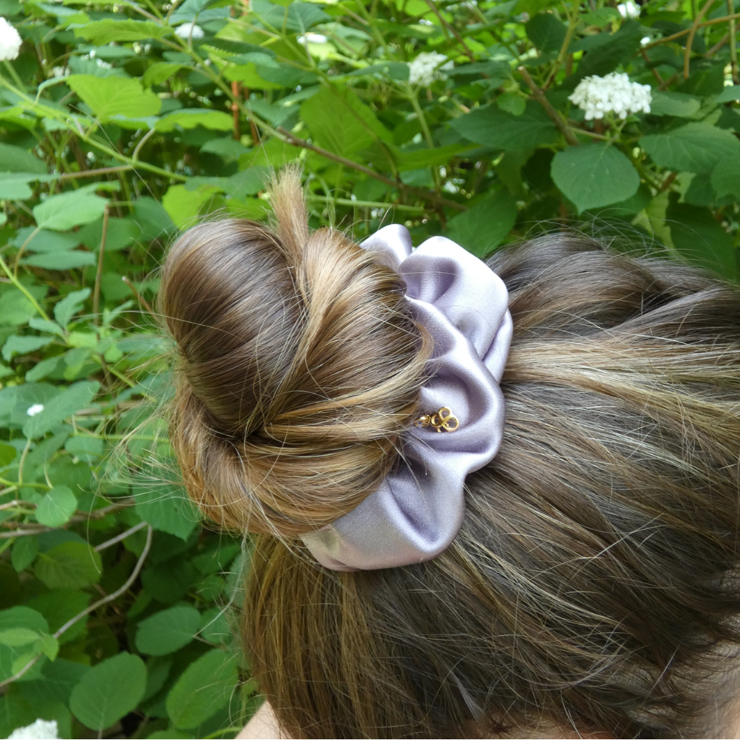 A lavender satin scrunchie with a small gold bead accent, holding up a brown and blonde high bun hairstyle outdoors against a background of green leaves and white flowers.