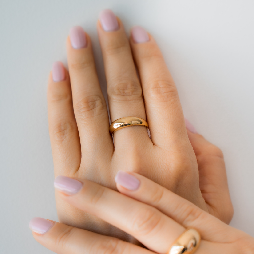 A hand wearing a gold dome ring with light pink nail polish on a neutral background.