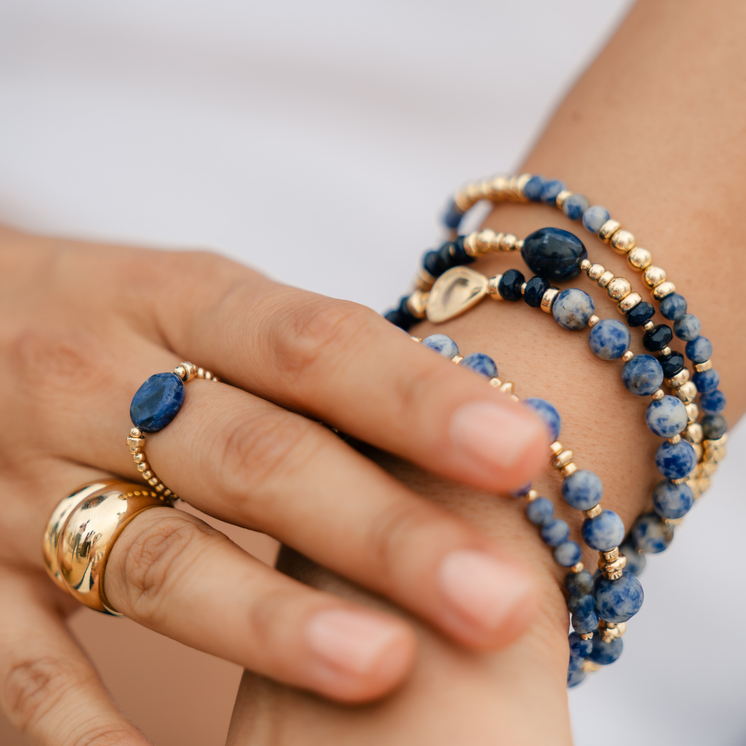 Hand wearing a stack of gold vermeil rings and blue sodalite beaded bracelets on a white background/ beblue bijoux