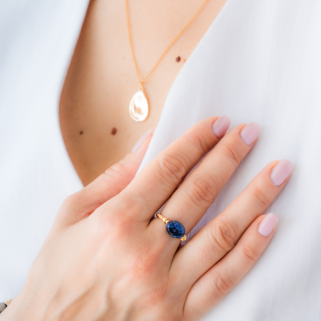 Hand wearing a blue sodalite gemstone elastic ring with a gold necklace charm / beblue bijoux