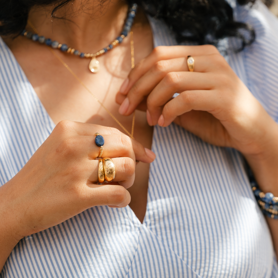 Close-up of a person wearing a stack of gold rings and a layering of necklaces with blue sodalite beads and pebble charm. / belue bijoux