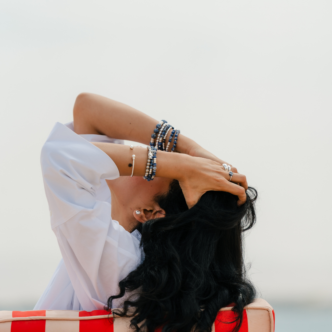 Woman with long black hair and a white top sitting on a red and white striped chair. She is wearing a silver  pebble earring with stacking rings and blue sodalite elastic bracelet .