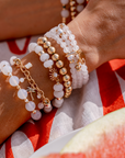 Close-up of a wrist wearing stacked beaded bracelets on a red and white checkered fabric background. The jewelry features white agate and gold accents, with palm tree and sun charms.
