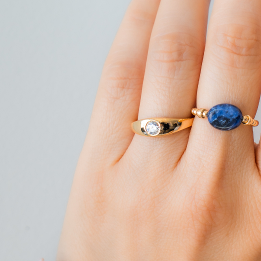 Hand wearing a gold ring with a zircon and an other elastic ring with a blue sodalite stone on a neutral background / beblue bijoux