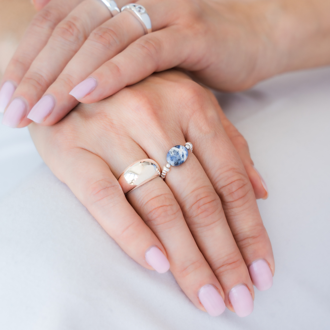 Hand wearing a stack of silver rings, including a blue sodalite elastic ring, on a white shirt. / beblue bijoux