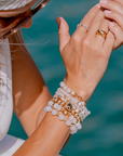 Close-up of a wrist wearing stacked beaded bracelets on a red and white checkered fabric background. The jewelry features white agate and gold accents, with palm tree and sun charms.
