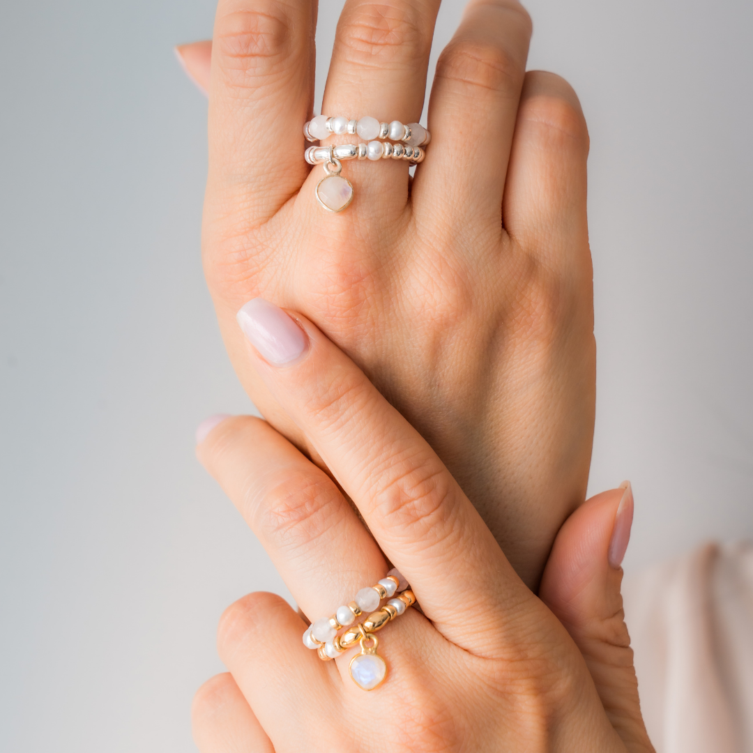 Close-up of a hand wearing multiple pearl rings and charm ring on a neutral background