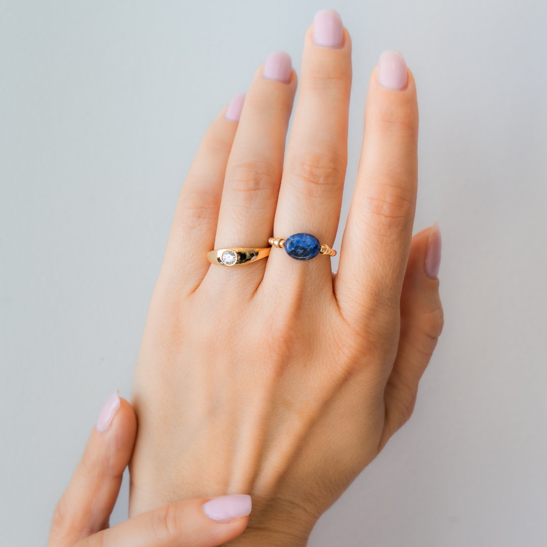 Hand wearing a stack of gold vermeil, including a blue sodalite elastic ring, on a white shirt. / beblue bijoux