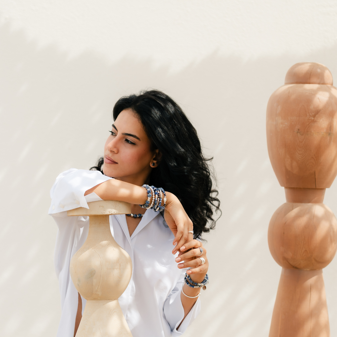 Woman in a white shirt leaning on a wooden mannequin against a plain background.She is wearing blue sodalite   elastic bracelets.