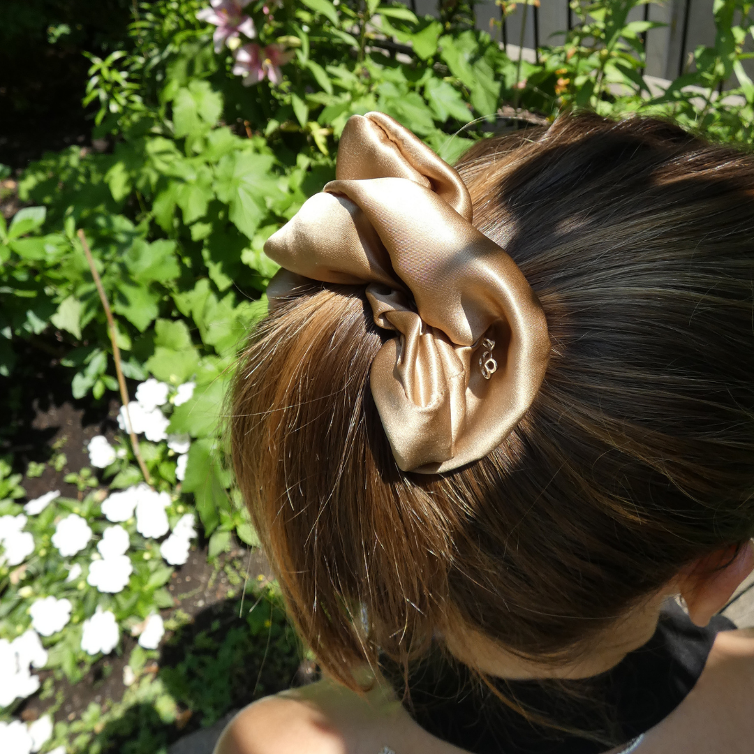 A large satin hair bow accessory in a champagne gold color, shown clipped to a woman's brown hair outdoors in a garden setting.