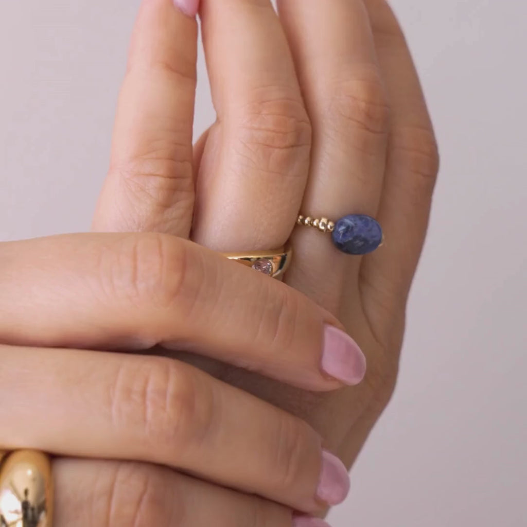 Hand wearing a stack of gold vermeil rings, including a blue sodalite elastic ring, on a white shirt. / beblue bijoux