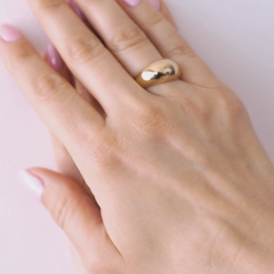 A hand wearing a gold dome ring with light pink nail polish on a neutral background.