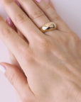 A hand wearing a gold dome ring with light pink nail polish on a neutral background.
